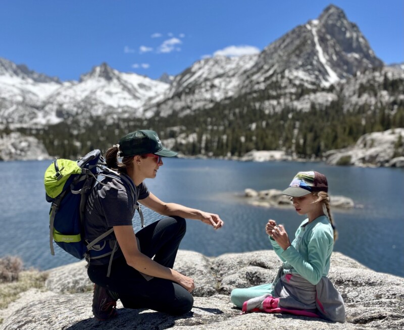 Two people wearing hiking gear sit on rocks by a lake with snow-capped mountains in the background. visit bishop