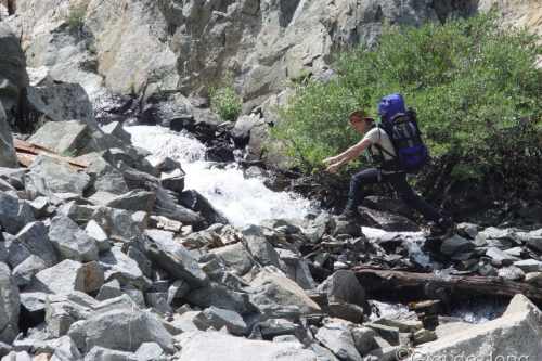 A hiker with a blue backpack carefully crosses a rocky stream in a mountainous area. visit bishop