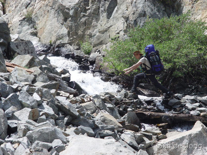 A hiker with a blue backpack carefully crosses a rocky stream in a mountainous area. visit bishop