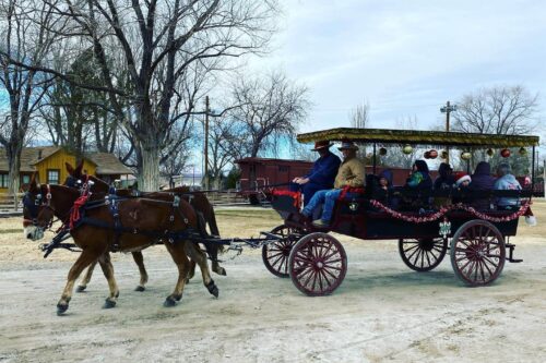 A horse-drawn carriage adorned with festive decorations is carrying a group of bundled-up people. The scene, reminiscent of Bishop, California, is set in a rustic area with bare trees and wooden buildings in the background, under a cloudy sky. visit bishop