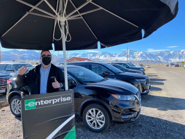 A person wearing a face mask and standing outdoors next to a sign that says "Enterprise" in Bishop, California. They are giving a thumbs-up gesture. Several black cars are parked beside them, and snow-capped mountains are visible in the background under a clear blue sky. visit bishop