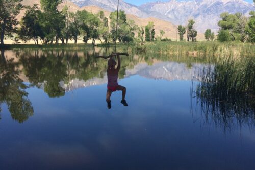 A person swings from a rope over a calm lake with trees and mountains in the background under the clear blue sky of Bishop, California. visit bishop