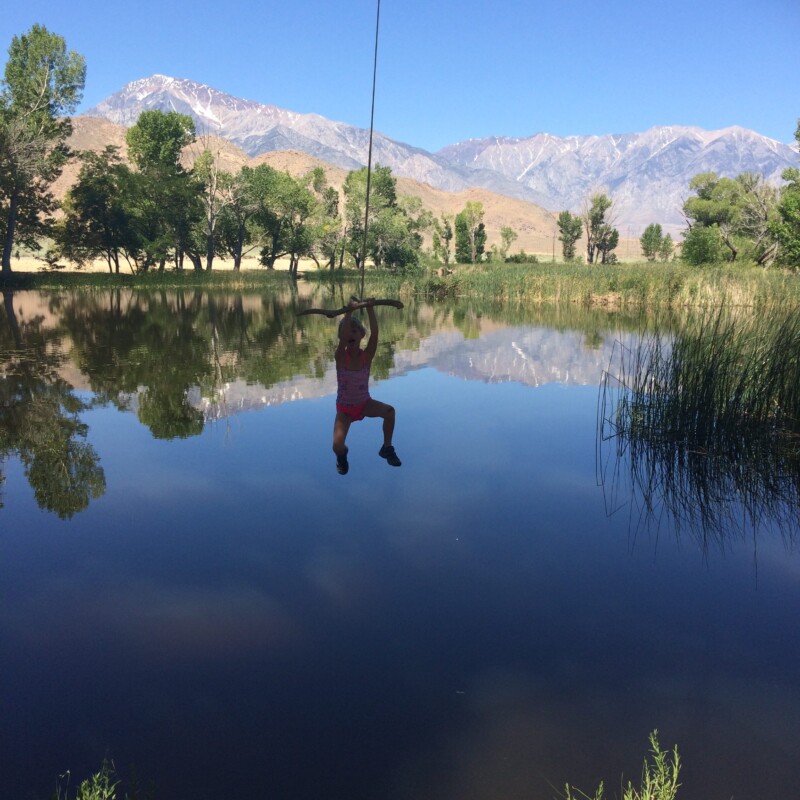 A person swings from a rope over a calm lake with trees and mountains in the background under the clear blue sky of Bishop, California. visit bishop