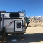 A person climbs the ladder on the side of a white and gray camper van in a desert landscape near Bishop, California, with rocky hills and snow-capped mountains in the background under a clear blue sky. visit bishop