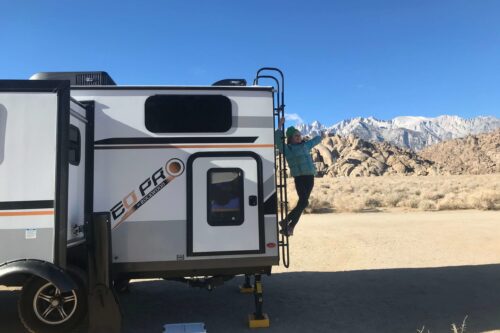 A person climbs the ladder on the side of a white and gray camper van in a desert landscape near Bishop, California, with rocky hills and snow-capped mountains in the background under a clear blue sky. visit bishop