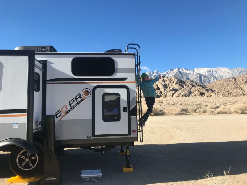 A person climbs the ladder on the side of a white and gray camper van in a desert landscape near Bishop, California, with rocky hills and snow-capped mountains in the background under a clear blue sky. visit bishop