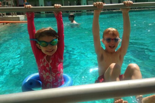 Two children in swimsuits wearing goggles and smiling at the camera hang from a poolside railing in Bishop, California. The girl on the left has a pink floral swimsuit and a blue pool noodle around her waist. The boy on the right wears red swim trunks. The water is bright blue. visit bishop