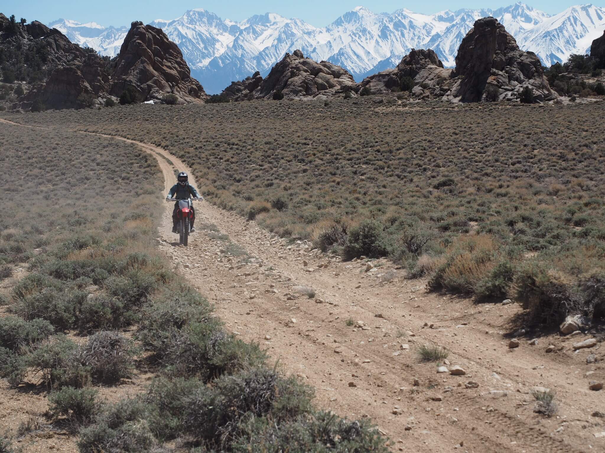 A person rides a motorcycle on a dirt path in the Eastern Sierra, navigating the mountainous, desert landscape near Bishop, California, with snow-capped peaks in the background. visit bishop