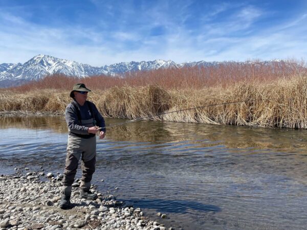 A person dressed in fishing gear is standing beside a stream surrounded by tall grasses in Bishop, California, holding a fishing rod. Snow-capped mountains are visible in the background under a partly cloudy sky. Pebbles and water form the foreground. visit bishop