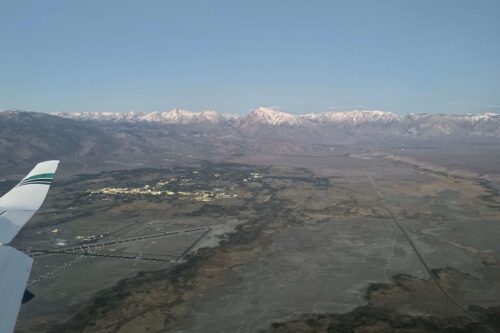 Aerial view from an airplane window showing a vast landscape with mountains and a town below during early evening. visit bishop