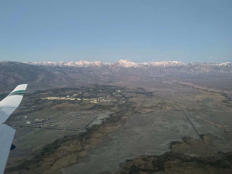 Aerial view from an airplane window showing a vast landscape with mountains and a town below during early evening. visit bishop