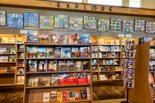 Bookshelf with various travel guides, maps, and nature guides, beneath a sign that reads "EXPLORE" and a colorful mural. visit bishop