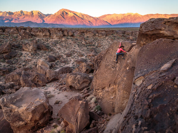 A lone climber wearing a pink jacket scales a large rock in the rugged desert landscape of Bishop, California, during sunrise. The sun casts a golden glow over distant mountain peaks, illuminating the rocky terrain with warm hues. visit bishop
