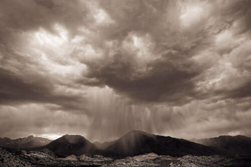 Dramatic black-and-white landscape showing rain falling over mountains beneath heavy clouds. visit bishop