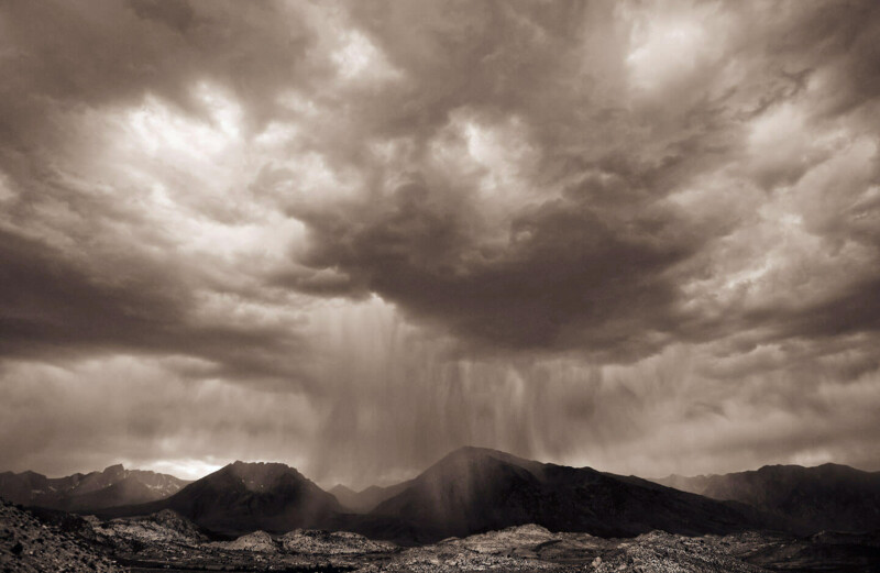 Dramatic black-and-white landscape showing rain falling over mountains beneath heavy clouds. visit bishop