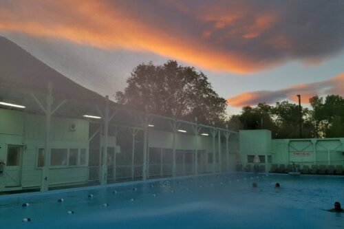 Outdoor pool at dusk with swimmers, buildings on either side, and a colorful sky with orange and purple clouds in the background. visit bishop