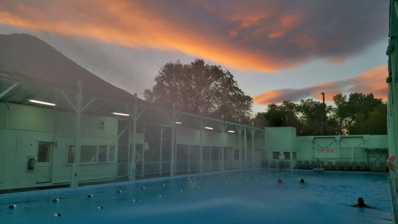 Outdoor pool at dusk with swimmers, buildings on either side, and a colorful sky with orange and purple clouds in the background. visit bishop