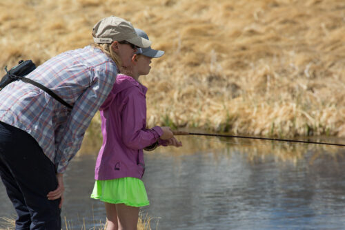 Two people, an adult and a child, are fishing by a calm body of water in Bishop, California. The child is wearing a bright green skirt, pink jacket, and hat, holding a fishing rod with the adult close by, offering guidance. The background features dry, yellow grasses. visit bishop