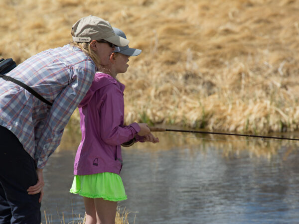 Two people, an adult and a child, are fishing by a calm body of water in Bishop, California. The child is wearing a bright green skirt, pink jacket, and hat, holding a fishing rod with the adult close by, offering guidance. The background features dry, yellow grasses. visit bishop