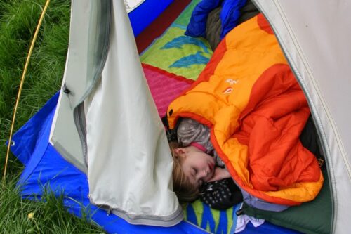 Child sleeping in an orange sleeping bag inside a tent on a colorful mat, with grass visible outside the tent in Bishop, California. visit bishop