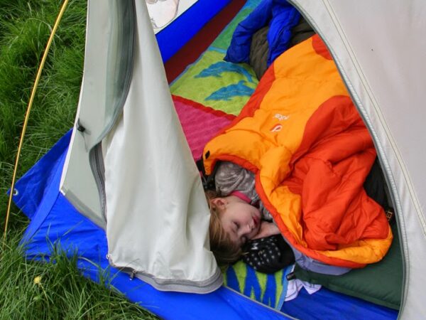 Child sleeping in an orange sleeping bag inside a tent on a colorful mat, with grass visible outside the tent in Bishop, California. visit bishop