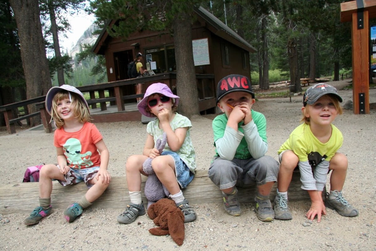 Four children in casual clothing and hats sit on a wooden bench outdoors, with a cabin in the background. visit bishop