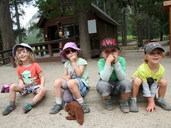 Four children in casual clothing and hats sit on a wooden bench outdoors, with a cabin in the background. visit bishop