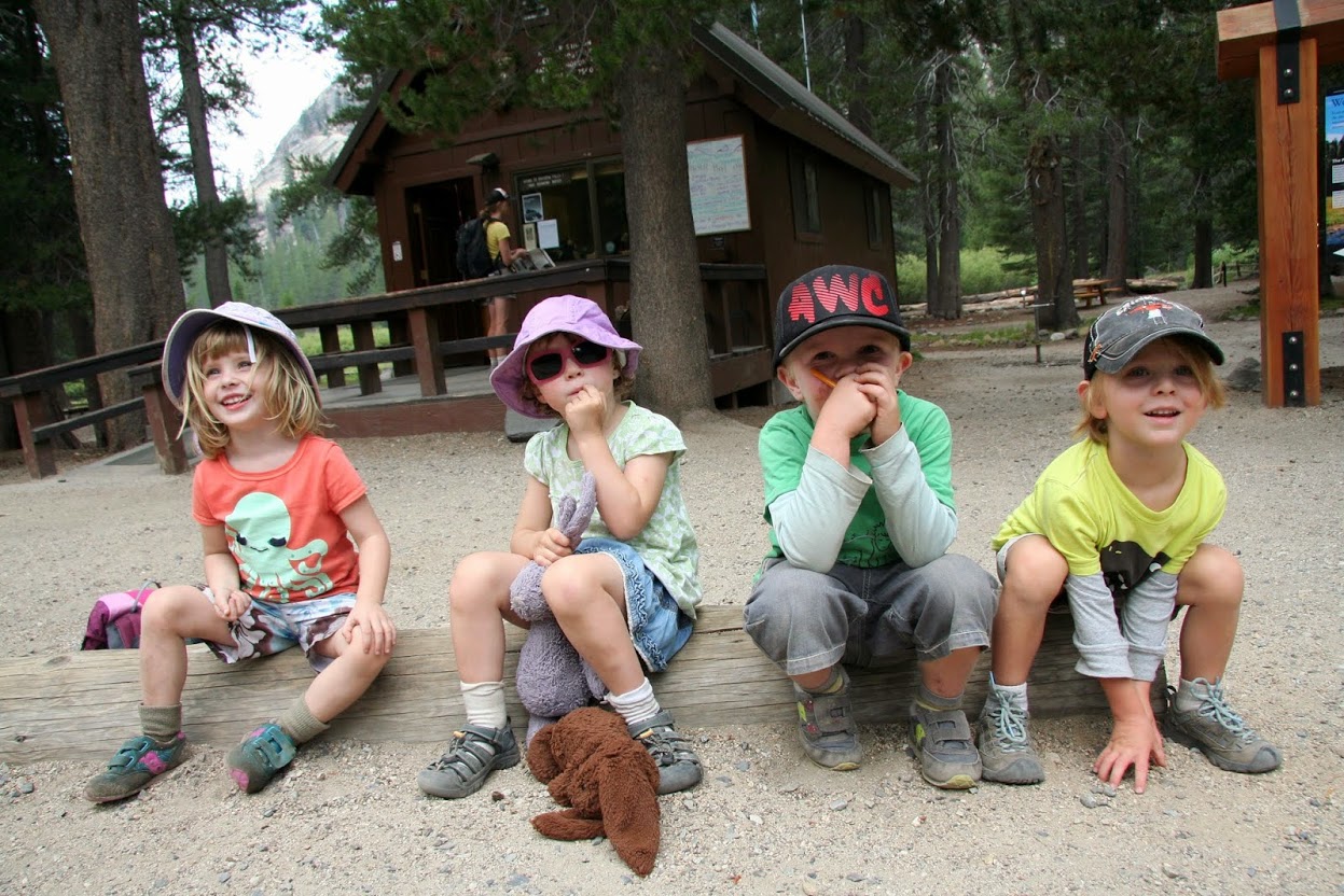 Four children in casual clothing and hats sit on a wooden bench outdoors, with a cabin in the background. visit bishop