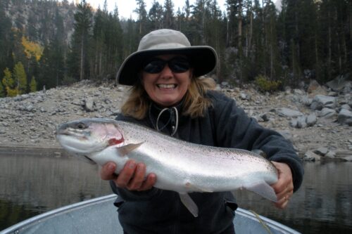 Smiling person in a hat holding a large fish on a boat with a forested rocky background. visit bishop