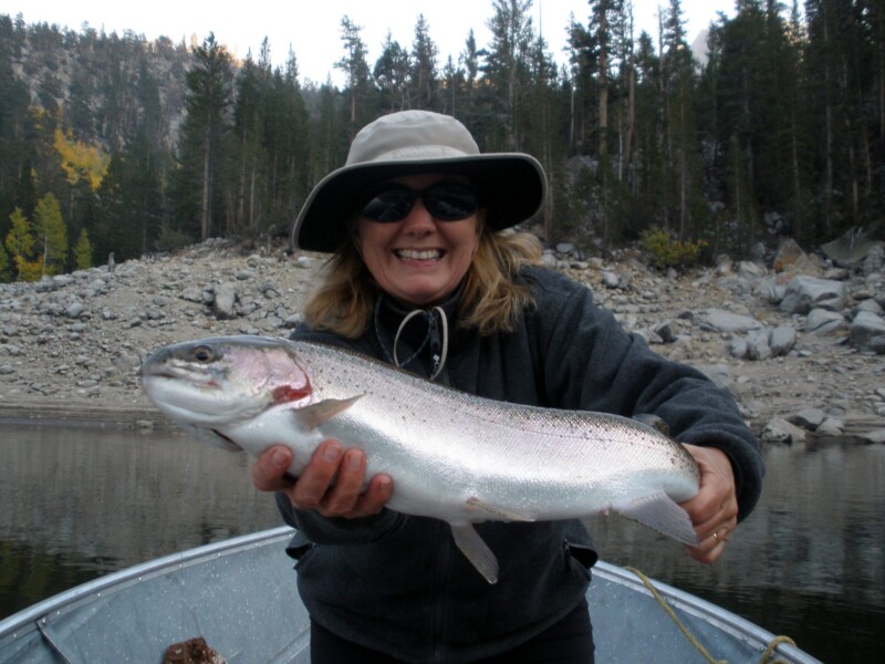 Smiling person in a hat holding a large fish on a boat with a forested rocky background. visit bishop