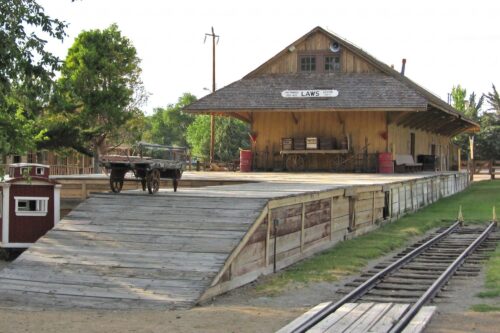 A historic wooden train depot with a platform, cart, and tracks in front of a building labeled "Laws. visit bishop