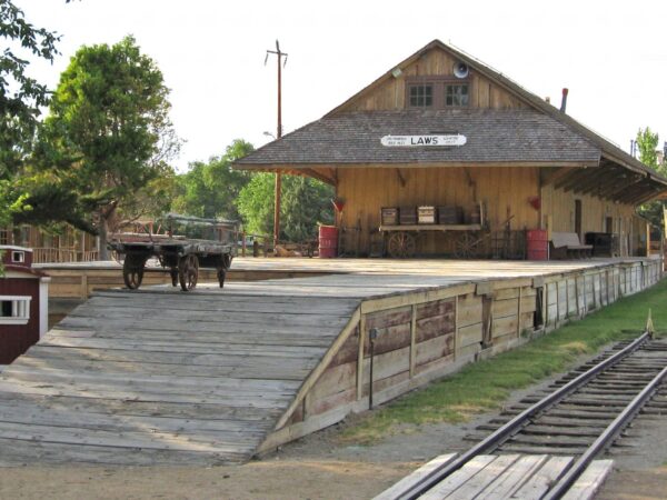 A historic wooden train depot with a platform, cart, and tracks in front of a building labeled "Laws. visit bishop