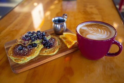 Pancakes with blueberries and a side of orange slices on a wooden board, next to a red cup of latte with foam art. visit bishop