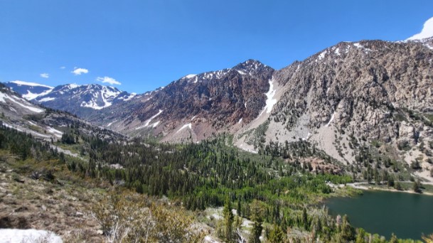 A panoramic view of a lush valley with green trees, a serene lake, and towering rocky mountains under a clear blue sky captures the breathtaking beauty of Bishop, California in the Eastern Sierra. visit bishop