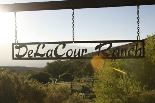 Sign reading "DeLaCour Ranch" hanging above a scenic view of hills and vegetation at sunset. visit bishop