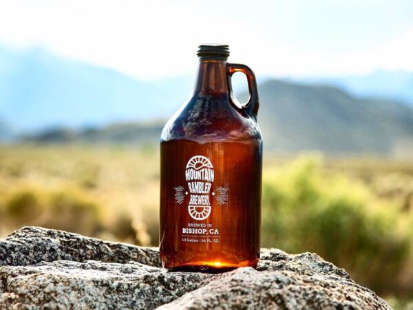 A brown glass jug with a Mountain Rambler Brewery label sits on a boulder in a mountainous landscape. visit bishop