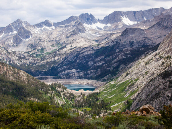 A mountainous landscape with rocky peaks, a small lake in the valley, and scattered greenery under a cloudy sky. visit bishop