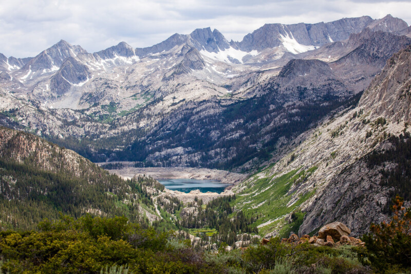 A mountainous landscape with rocky peaks, a small lake in the valley, and scattered greenery under a cloudy sky. visit bishop