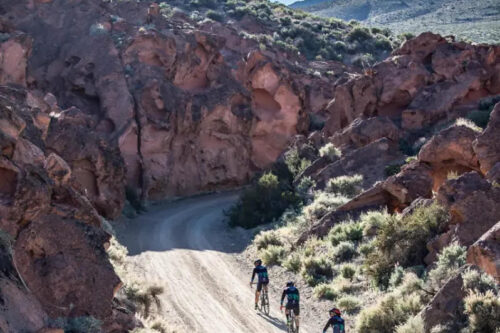 Three cyclists ride along a dirt road through a rocky canyon landscape under a clear blue sky. visit bishop