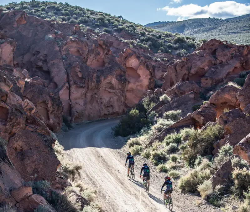 Three cyclists ride along a dirt road through a rocky canyon landscape under a clear blue sky. visit bishop