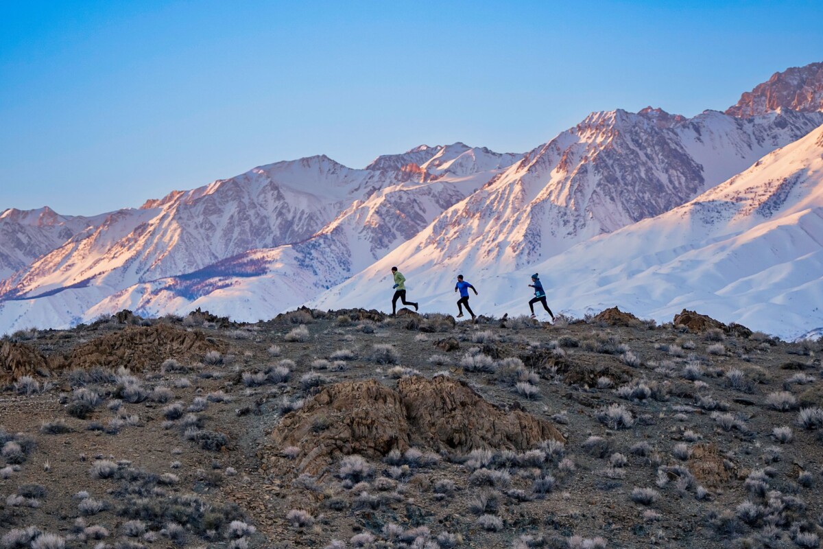 Three people running on a mountainous terrain with snow-capped peaks and a clear blue sky in the background. visit bishop