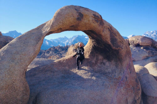 Person sits under a rock arch in a mountainous desert landscape with clear blue sky. Snowy peaks seen in the background. visit bishop