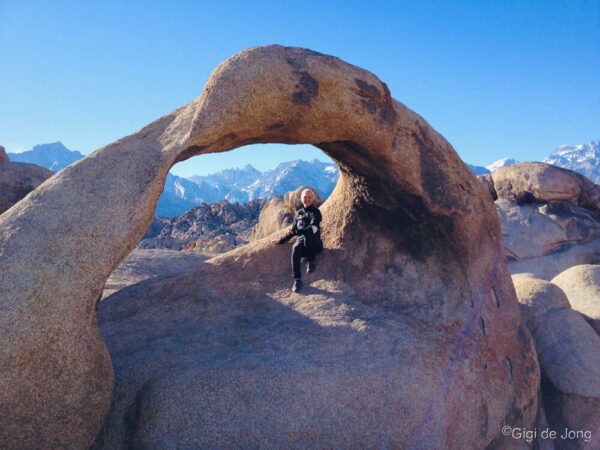 Person sits under a rock arch in a mountainous desert landscape with clear blue sky. Snowy peaks seen in the background. visit bishop