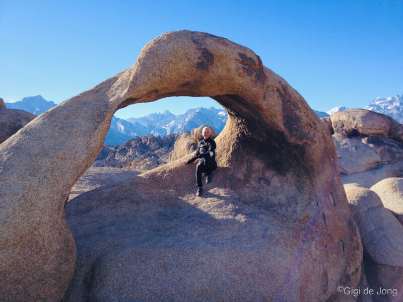 Person sits under a rock arch in a mountainous desert landscape with clear blue sky. Snowy peaks seen in the background. visit bishop