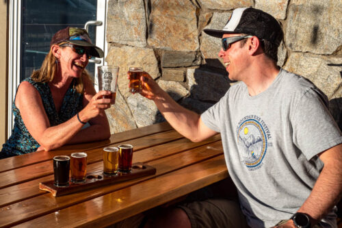 Two people sit at a wooden outdoor table in Bishop, California, laughing and clinking glasses of beer. A flight of beer samples is in front of them. The woman wears a sleeveless top and cap, the man a T-shirt, shorts, cap, and sunglasses. Stone wall and window are in the background. visit bishop