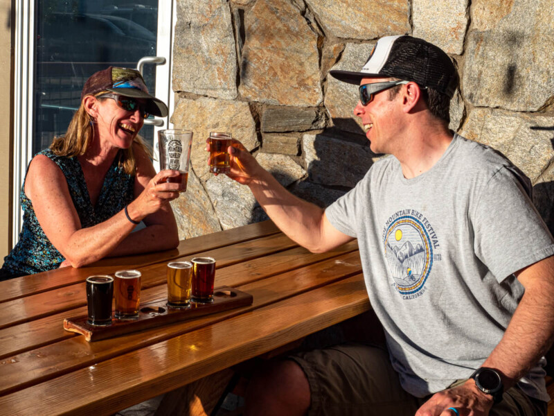 Two people sit at a wooden outdoor table in Bishop, California, laughing and clinking glasses of beer. A flight of beer samples is in front of them. The woman wears a sleeveless top and cap, the man a T-shirt, shorts, cap, and sunglasses. Stone wall and window are in the background. visit bishop