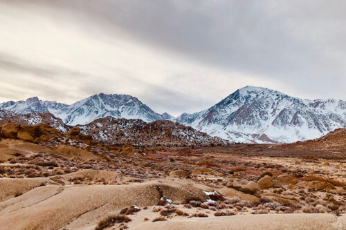 A vast desert landscape with sparse vegetation, leading to rugged, rocky hills in the foreground. Snow-capped mountain peaks rise majestically in the background under an overcast sky, capturing the raw beauty of Bishop, California. The photo is by Gigi de Jong. visit bishop