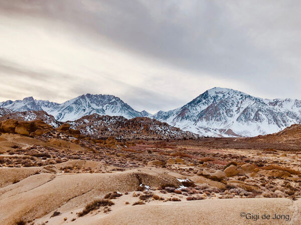 A vast desert landscape with sparse vegetation, leading to rugged, rocky hills in the foreground. Snow-capped mountain peaks rise majestically in the background under an overcast sky, capturing the raw beauty of Bishop, California. The photo is by Gigi de Jong. visit bishop