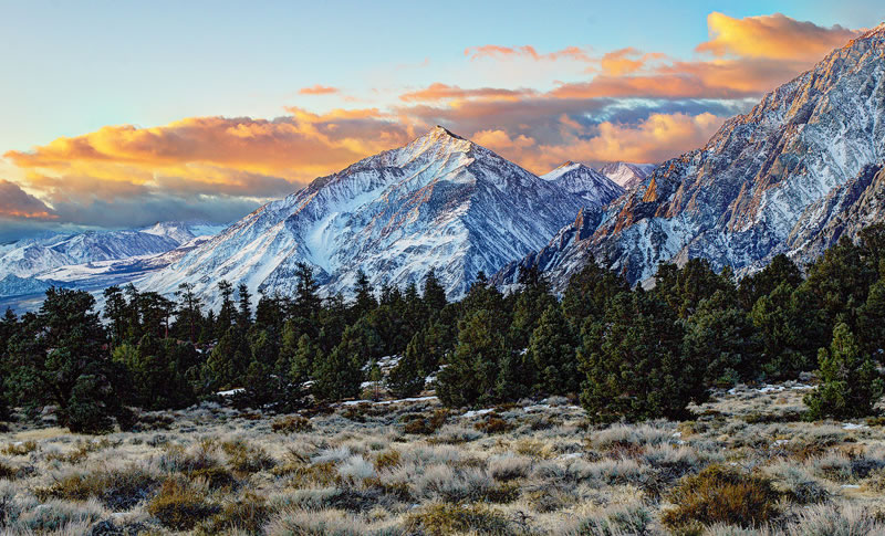 Snow-capped mountain range at sunset with a forested foreground and colorful clouds in the sky. visit bishop