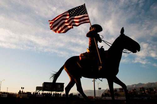 Silhouette of a person on horseback holding an American flag against a dusk sky in a rodeo arena. visit bishop